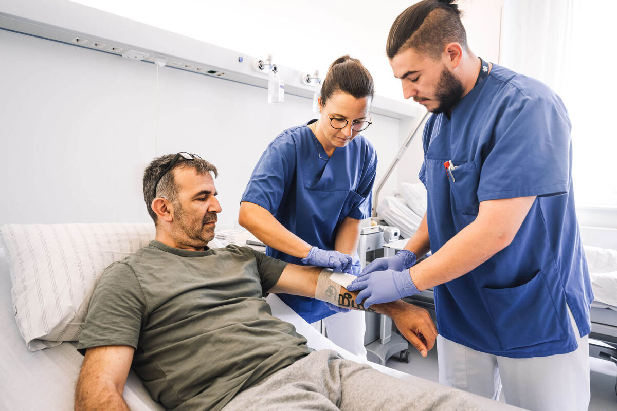 Das Bild zeigt eine pflegerische Mitarbeiterin und eine männliche Pflegefachkraft bei der Behandlung eines im Bett liegenden Patienten.